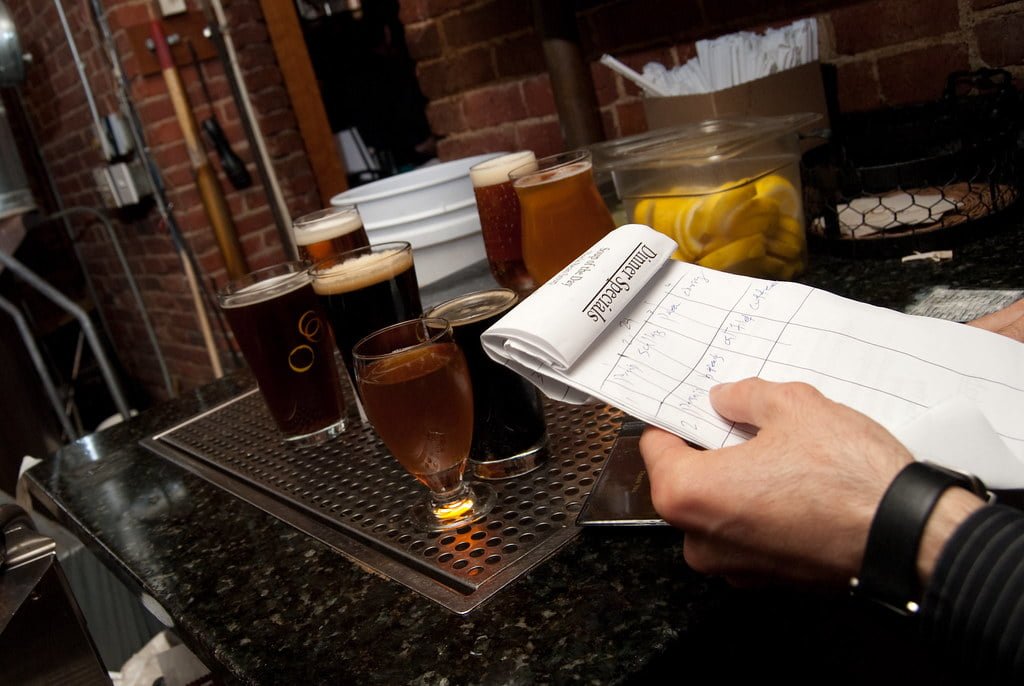 A person's hand holding a notepad on a bar counter with various glasses of beer and a container of lemon slices in the background.