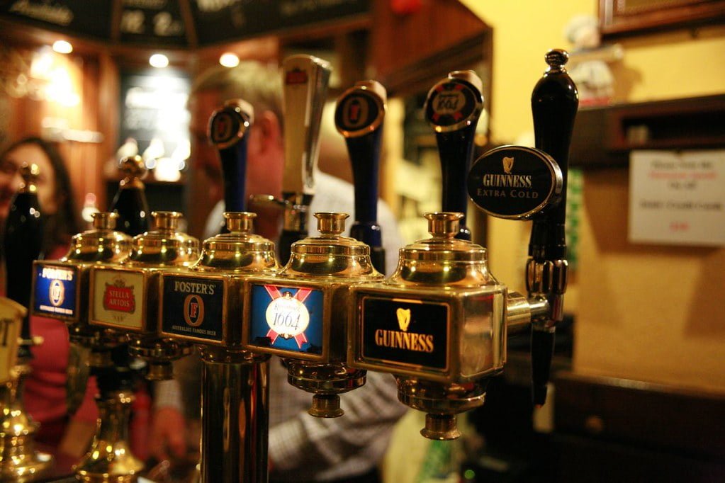 A row of beer taps with various brand labels, including Foster's, Stella Artois, and Guinness, in a busy pub with patrons in the background.