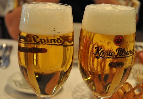 Two glasses of König Pilsener beer with frothy heads on a table, with a basket and tableware in the background.