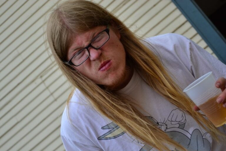 A person with long blonde hair and glasses making a face of distaste while holding a plastic cup.