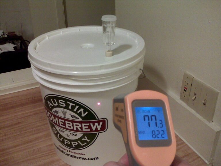 A homebrew fermentation bucket from Austin Homebrew Supply with an airlock on the lid, and a digital thermometer displaying a temperature of 77.3 degrees Fahrenheit, with a maximum recorded temperature of 82.2 degrees.
