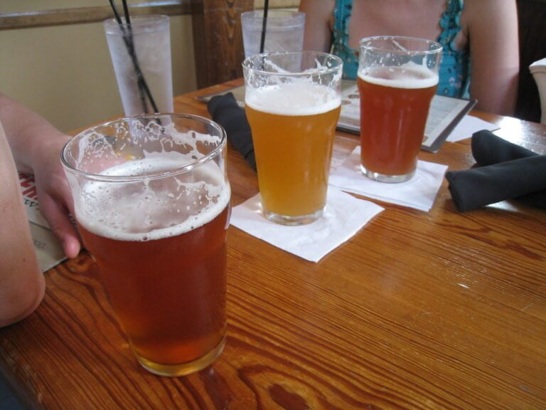 Three glasses of beer on a wooden table with a person's arm partially visible on the left and another person across the table; two drinks with straws and napkins also on the table.