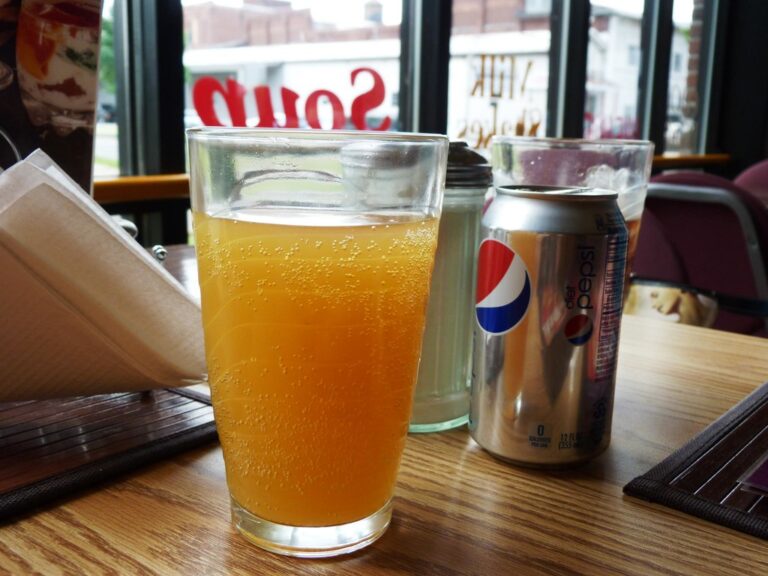 A glass of orange juice on a wooden tabletop with a can of Diet Pepsi and a napkin dispenser, with a window showing the reversed signage of a Sonic Drive-In in the background.