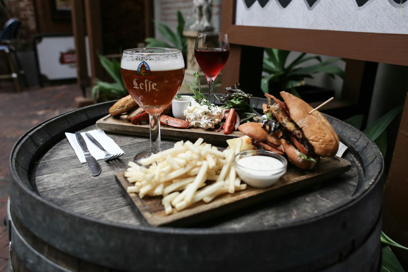 A platter of food on a wooden barrel table, featuring a glass of beer, red wine, fries with dipping sauce, a sandwich, salad, and various grilled items. Utensils and a napkin are alongside the plate.