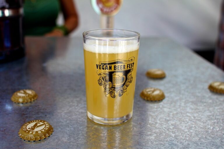 A glass of beer with the text "VEGAN BEER FEST" on it, placed on a metal table with scattered bottle caps, with a blurred background featuring a person.