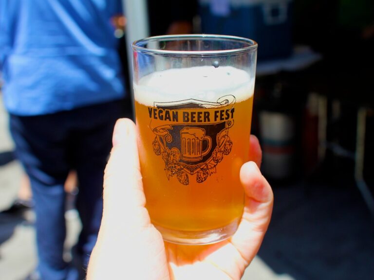 A hand holding a half-full glass of beer with the text "VEGAN BEER FEST" on it, with a blurred background of a crowd.