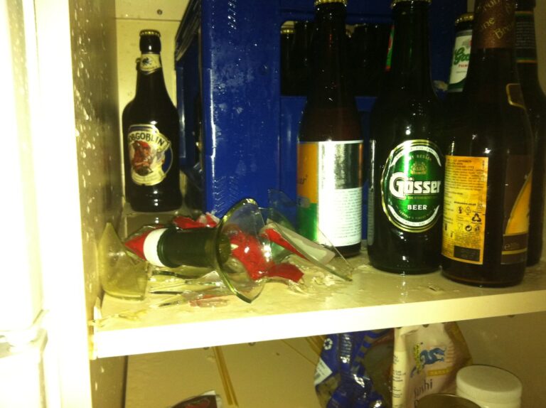 A broken beer bottle with liquid spilled on a shelf alongside several other unopened beer bottles.