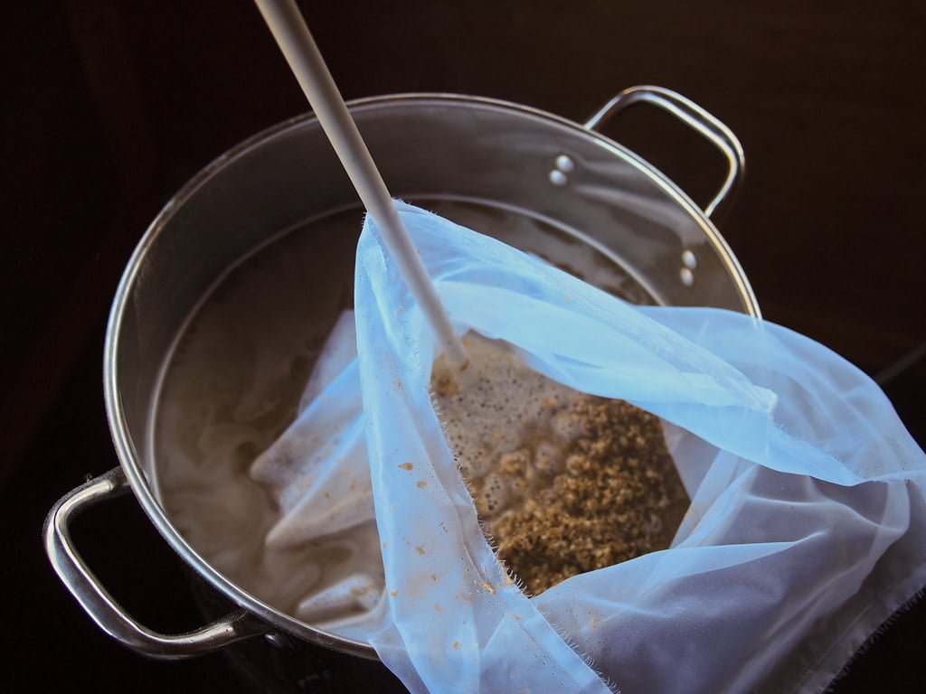 A stainless steel pot with a plastic brewing bag containing grains, immersed in liquid, likely during the beer brewing process.