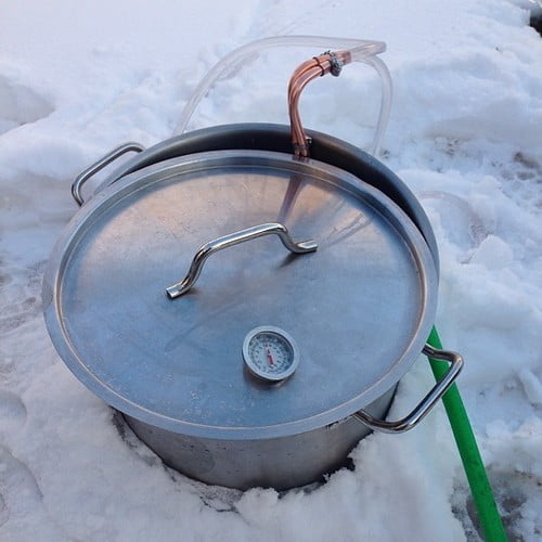 A metal brewing kettle with a thermometer and copper tubing, sitting on snow.