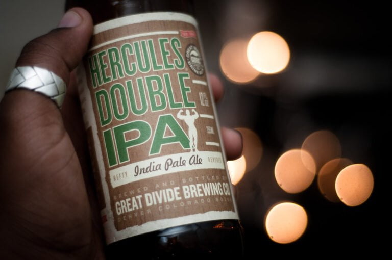 A close-up of a person's hand holding a Hercules Double IPA bottle from Great Divide Brewing Co., with soft bokeh lights in the background.