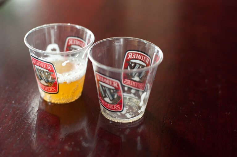 Two plastic sampling cups with Widmer Brothers Brewery logos, one half full with beer and the other empty, on a wooden table surface.