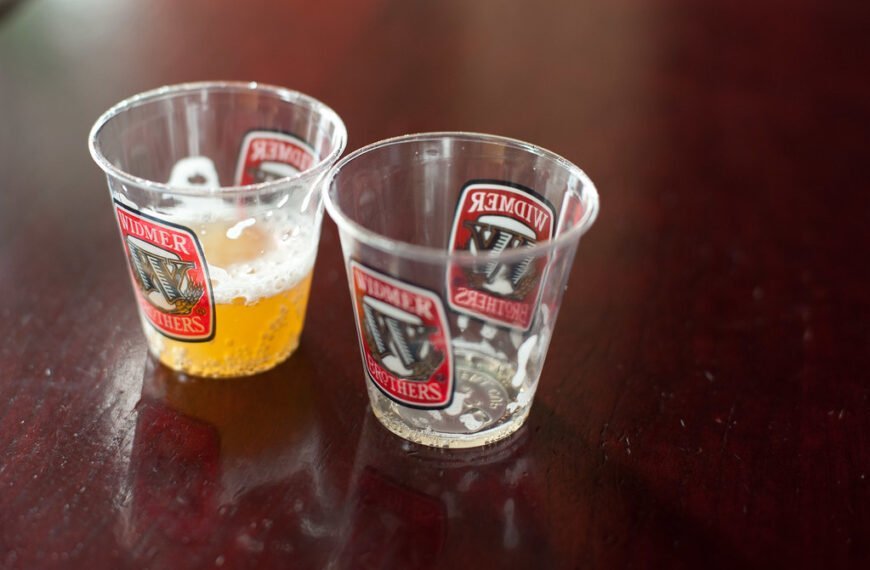 Two plastic sampling cups with Widmer Brothers Brewery logos, one half full with beer and the other empty, on a wooden table surface.
