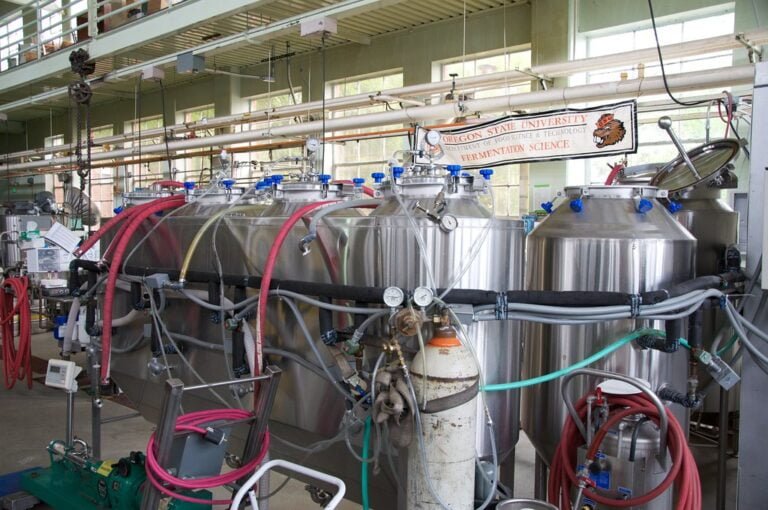 A laboratory equipped with multiple stainless steel fermentation tanks connected by a network of hoses and pipes, with signage indicating it belongs to the Oregon State University Department of Food Science and Technology Fermentation Science program.