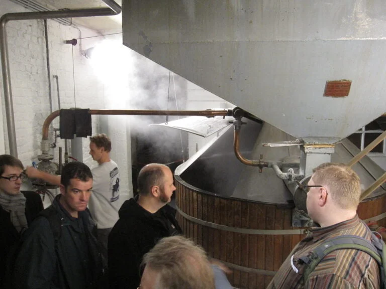 A group of people touring an industrial facility with large brewing tanks and steam.