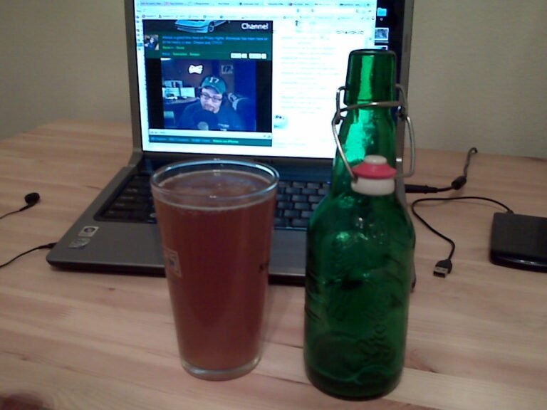 A laptop on a wooden desk displaying a webpage with a person in front of a computer, next to a glass filled with amber liquid and a green bottle with a flip-top cap.