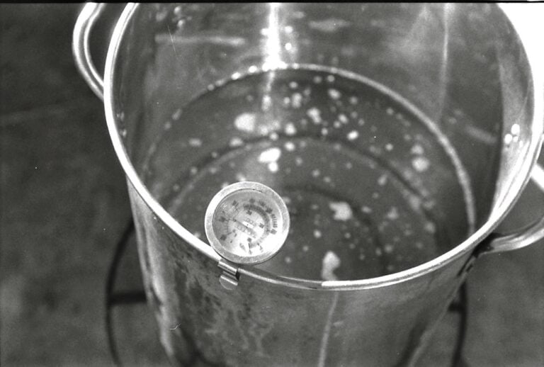 A black and white photo of a stainless steel cooking pot with water and a thermometer clipped to the side.