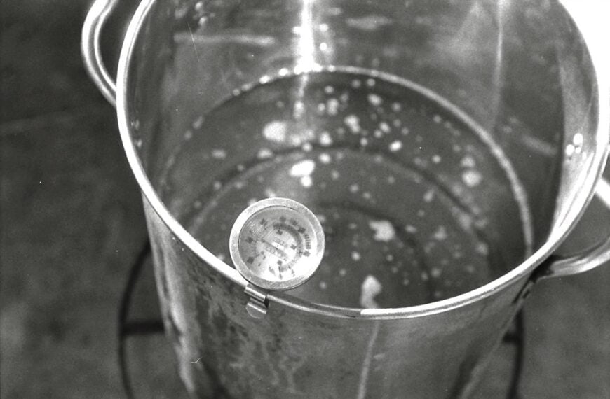 A black and white photo of a stainless steel cooking pot with water and a thermometer clipped to the side.