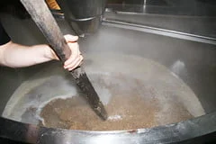 A person's hands are stirring a large metal brewing vat with a paddle, where brewing mash is visible.