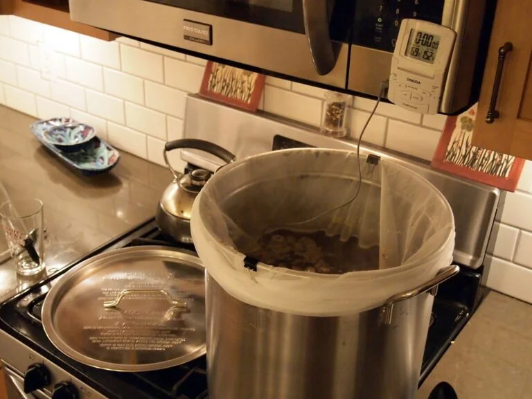 A home brewing setup in a kitchen with a large metal pot on the stove, displaying a bag filled with grains inside the pot, a digital timer hanging nearby, and various kitchen utensils and items in the background.
