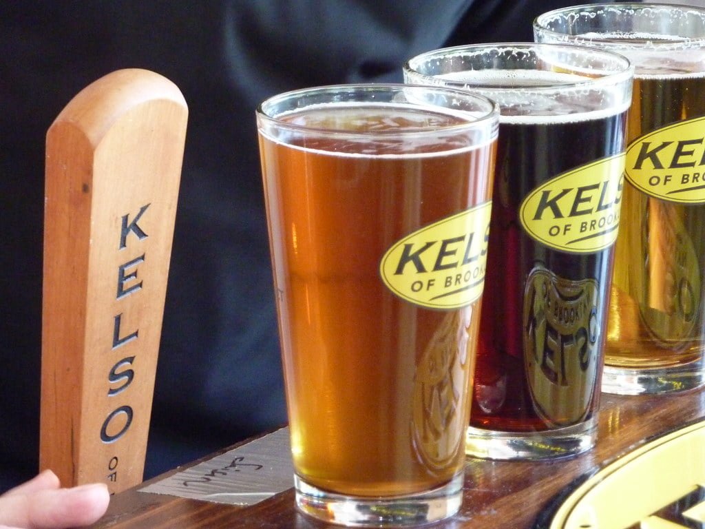 Three pints of beer with varying colors from pale amber to dark brown are lined up next to a wooden tap handle with the word "KELSO" on it. Each glass has a logo that reads "KELSO of Brooklyn." The background is indistinct.