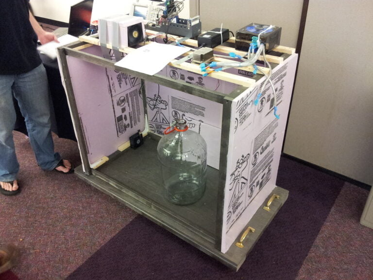 A homemade booth constructed from foam insulation panels covered with diagrams and notes, featuring various electronic components on its surface and a large empty glass carboy beside it. A person stands partially visible at the left side of the booth, with their hand on a piece of paper.