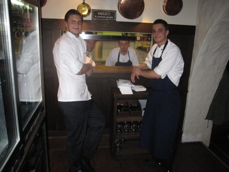 Three male kitchen staff posing in what appears to be a restaurant kitchen, with one behind a service window and two standing in front of it, all wearing chefs' uniforms. A sign reading "SMILE! It confuses people" is visible above the service window.