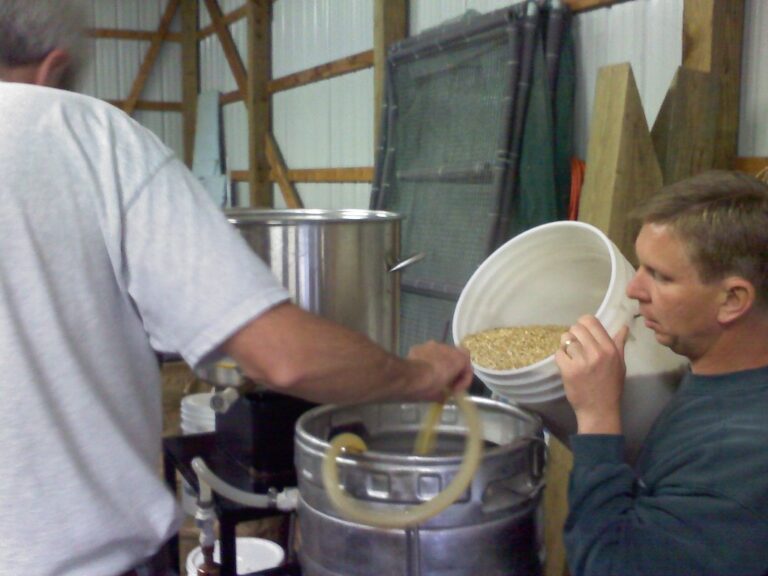 Two men working with brewing equipment in a workshop, with one adding grains into a brewing kettle.