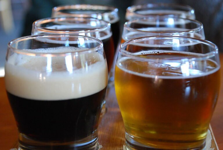 Six glasses of beer on a table with varying colors and head of foam, showcasing different beer types.