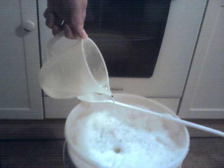 A person pouring a clear liquid from a jug into a bowl of frothy mixture on a kitchen floor.