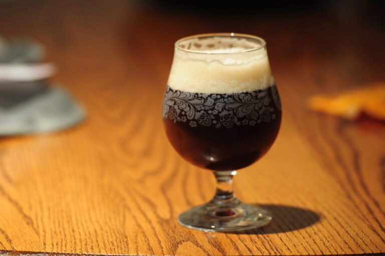 A glass of dark beer with a frothy head placed on a wooden surface with soft-focused background items.