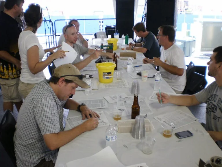 A group of people sitting at a long table with papers and pens, some are discussing while others are focused on writing, in a well-lit indoor setting with drinks and water bottles scattered on the table.
