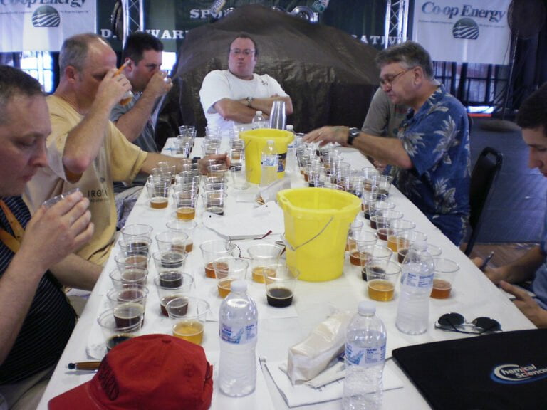 Men sitting at a table with multiple beer glasses for tasting, with one man taking notes and others focusing on their samples.