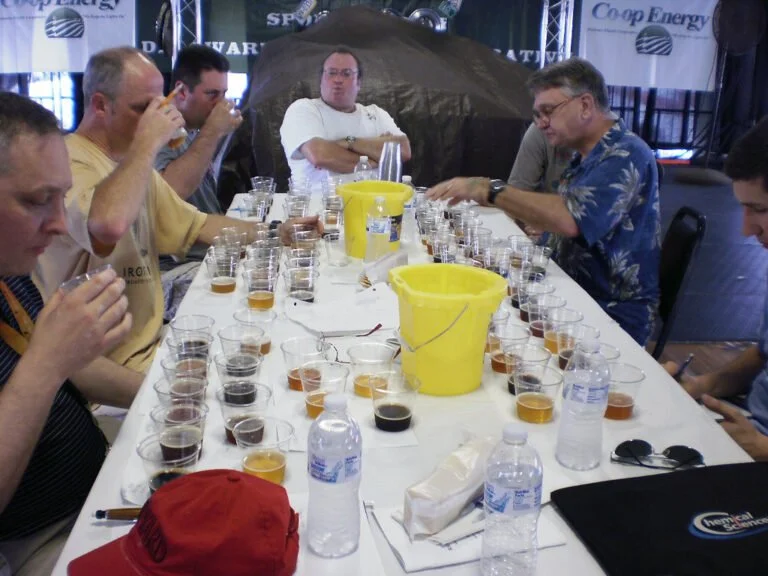 Men sitting at a table with multiple beer glasses for tasting, with one man taking notes and others focusing on their samples.
