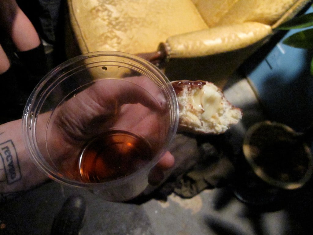 A person holding a clear plastic cup with a dark amber liquid, and a partially eaten pastry with white filling, in a dimly lit room.