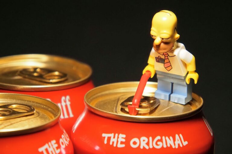 A Lego figure resembling Homer Simpson using a crowbar to open a beer can on a black background, with more cans in the background.