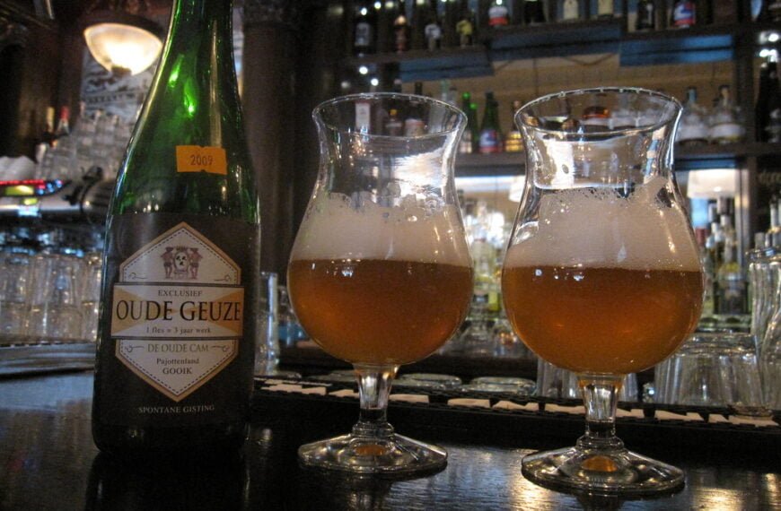 Alt text: A bottle labeled "Oude Geuze" dated 2009 with two tulip glasses of amber beer, on the counter of a bar with a backdrop of various bottles.