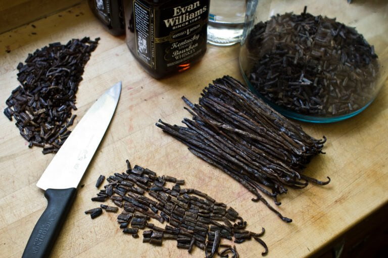 A kitchen countertop with chopped and whole vanilla beans, a chef's knife, and a bottle of whiskey.