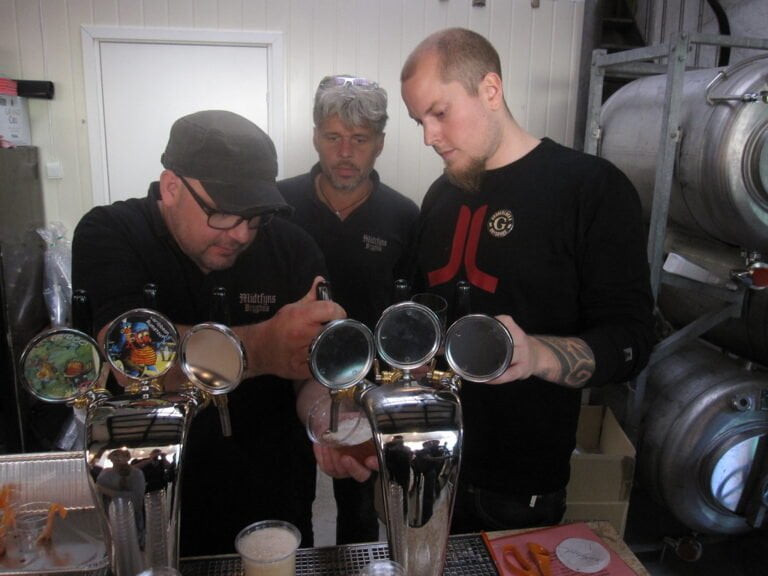 Three men are standing behind a bar with beer taps, one of whom is pouring a pint of beer while the others watch. There's brewing equipment in the background.