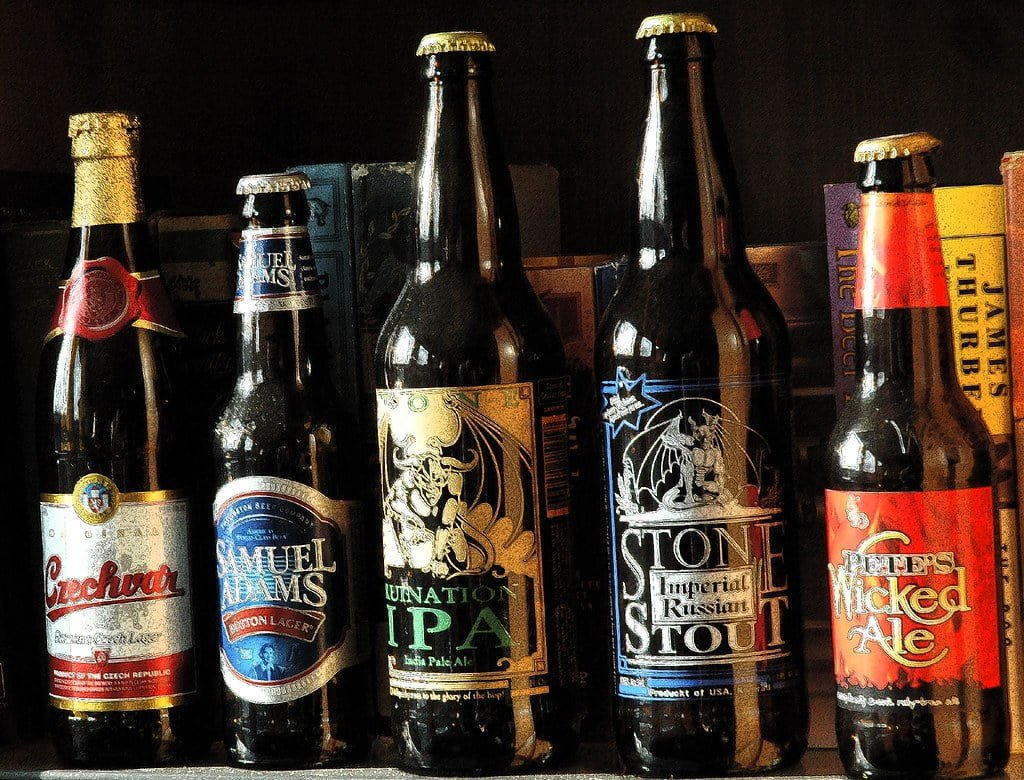 A collection of various beer bottles with different labels, including brands like Samuel Adams and Stone Imperial Stout, displayed in a row.