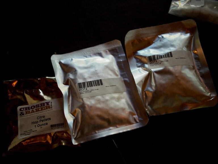 Three sealed packages of hop pellets for brewing on a dark surface, with labels indicating the variety as "Citra" and different alpha acid percentages.