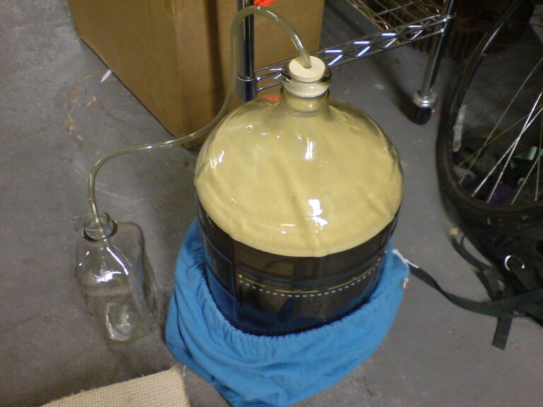 A carboy covered with a blue cloth in a fermentation setup, with an airlock attached and a clear tube running into a water-filled jar, placed on a concrete floor in a storage area.
