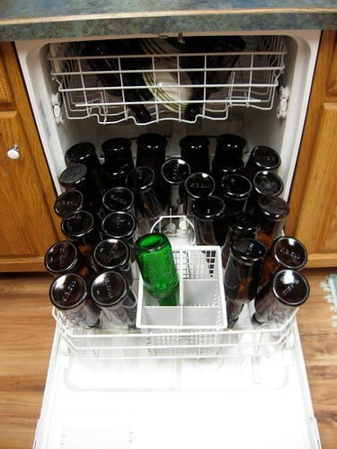 Open dishwasher filled with upside-down black bottles and one green amidst them, set in a kitchen with wooden cabinets.