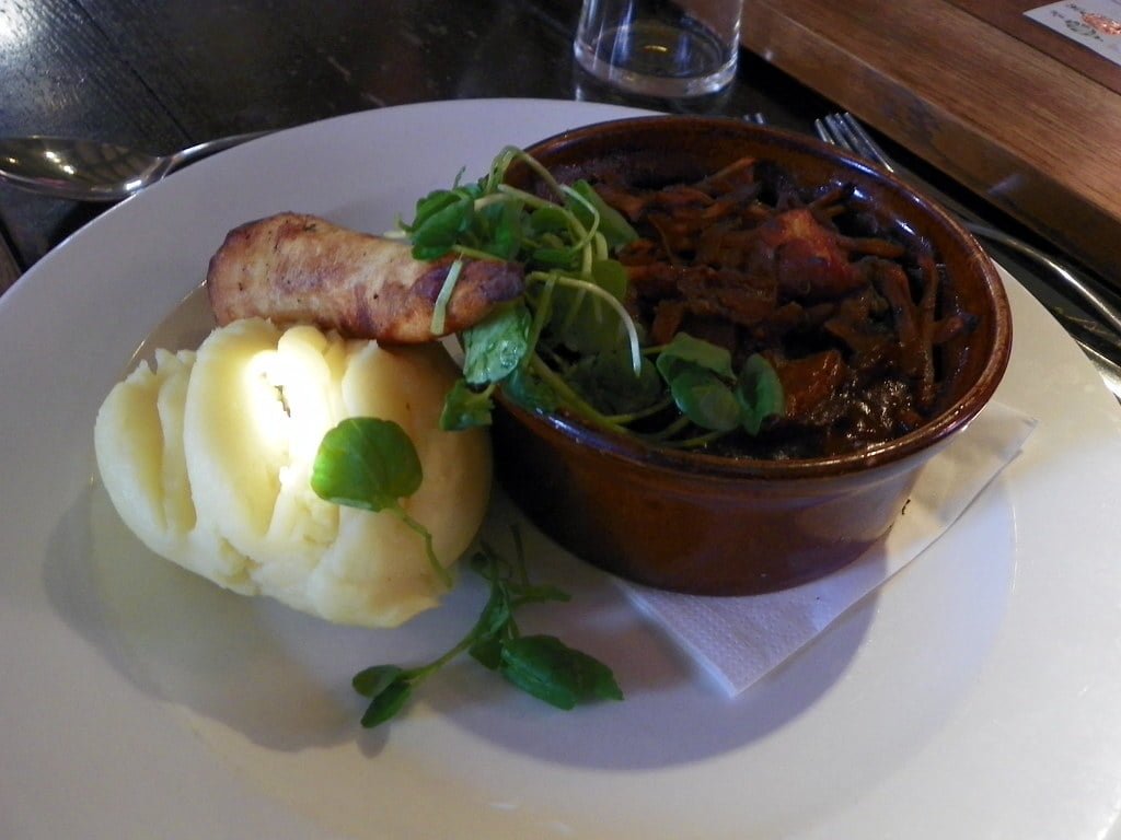 A plate with mashed potatoes and a sprig of greens beside a bowl of stew, set on a wooden table.