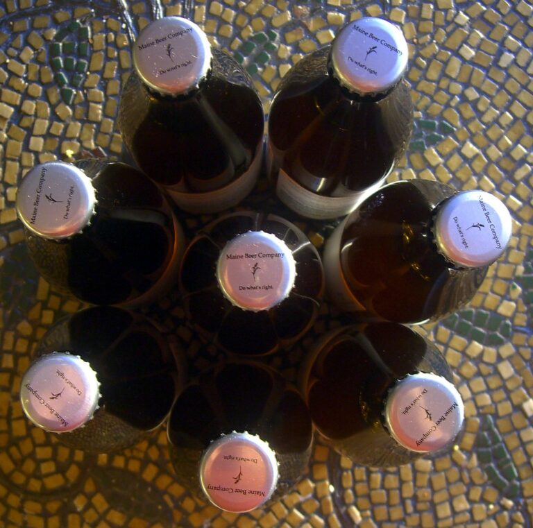 Nine bottles of beer from Maine Beer Company arranged in a circular pattern on a mosaic table, viewed from above.