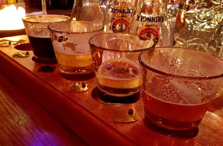 Five variously filled beer glasses on a wooden bar top, exhibiting a range of colors from dark to light, with some froth visible on the sides of the glasses. Bottles are visible in the background with a warm, dimly lit ambiance.