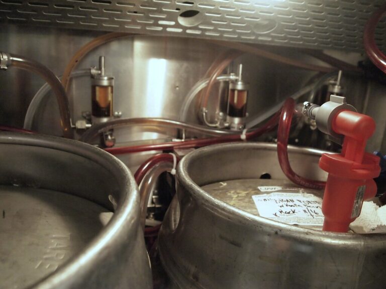 Interior of a brewery showing stainless steel kegs connected by red hoses and pressure gauges, with a handwritten note visible on one of the kegs.