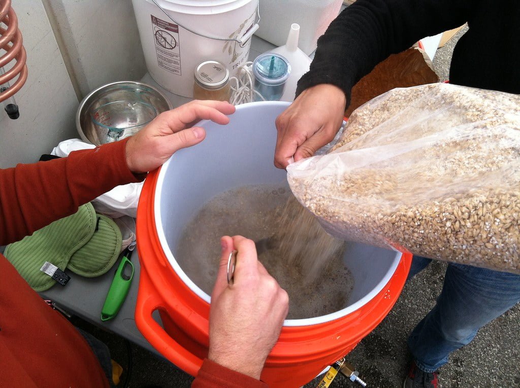 Two individuals are pouring grains from a clear plastic bag into a large white and orange bucket, likely engaging in a homebrewing process. The setup includes a metal bowl, scissors, and other brewing tools on a table.