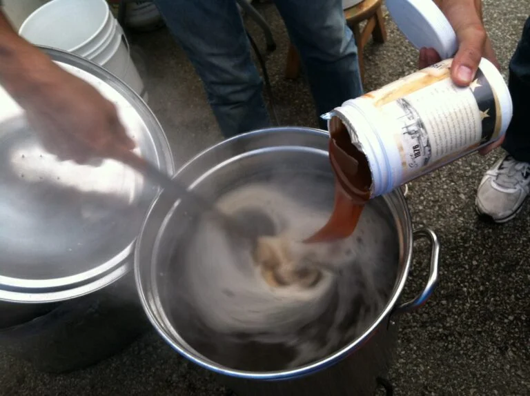 A person pouring a liquid from a paper container into a large metal pot with swirling contents, next to other pots and containers on an asphalt surface, suggesting an outdoor cooking or brewing activity.