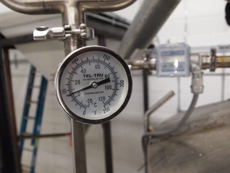 Analog industrial thermometer attached to a metallic pipe with a blurred background of industrial equipment.
