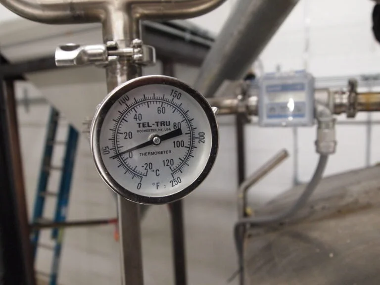 Analog industrial thermometer attached to a metallic pipe with a blurred background of industrial equipment.